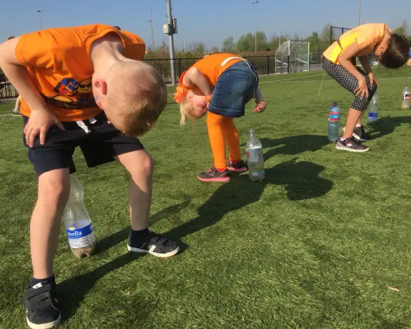 Barn i orange kläder böjer sig framåt för att försöka fånga en flaska med munnen. En del av kalas lekarna.