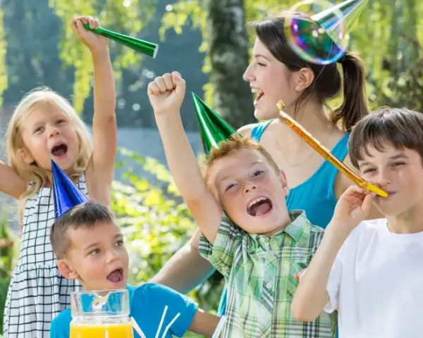 Barn firar ett 7-årskalas utomhus med ballonger, serpentiner och såpbubblor. Glada barn med partyhattar blåser i tutor.