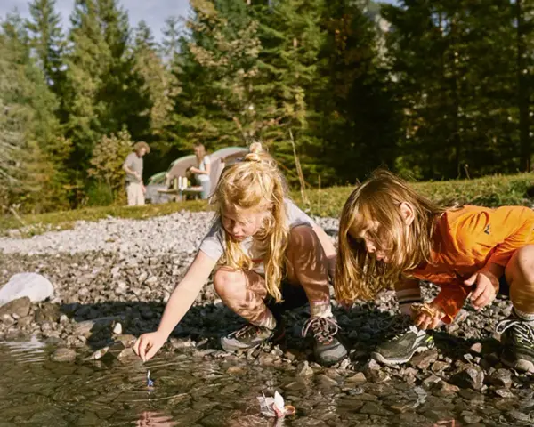 Två barn leker med pappersbåtar vid en sjö, kanske en del av roliga femkampsgrenar. I bakgrunden syns ett tält och träd.
