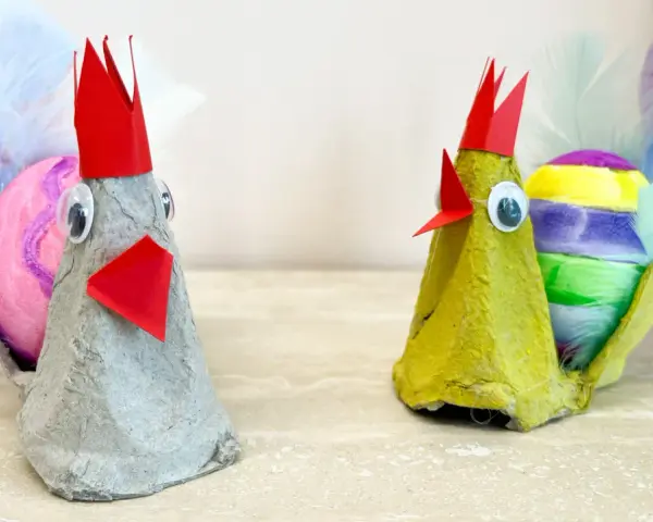 Två påskkycklingar gjorda av äggkartonger, med färgglada ägg och fjädrar. Ett roligt pyssel för påsk barn.