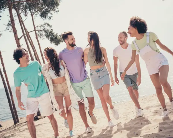 En grupp glada vänner på stranden, en riktig tipspromenad med twist. De skrattar och går längs vattnet bland tallar.