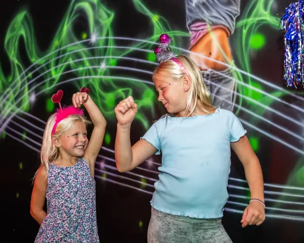 Två glada barn med diadem har roliga lekar inomhus. De dansar framför en neonfärgad bakgrund.