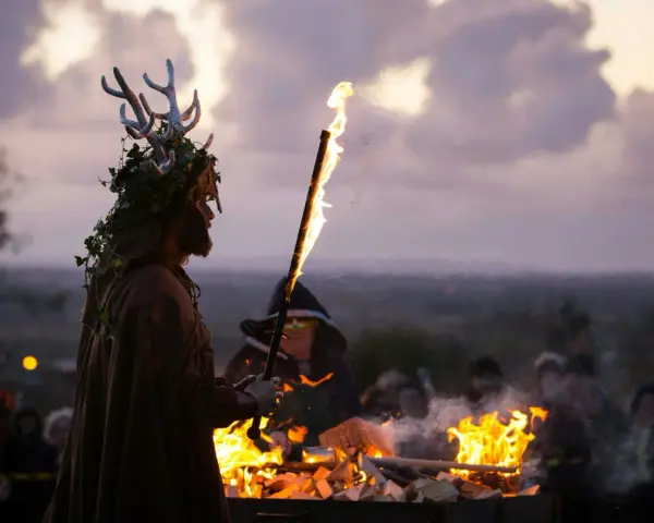 En person med hjorthorn och grönska på huvudet håller en fackla vid en eld. En annan person i mörk klädnad står bredvid. En del av en samling med halloween fakta.