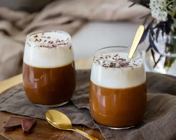 Två glas med krämig kaffe Karlsson, toppad med vispad grädde och chokladströssel. En guldig sked ligger i det ena glaset.