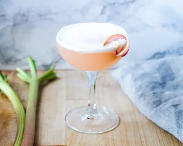 En rosa rabarber sour-cocktail med skum och en rabarberdekoration. Två rabarberstjälkar ligger bredvid.