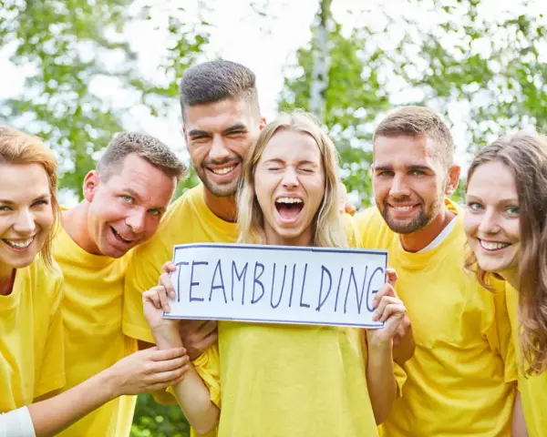 En grupp glada människor i gula tröjor håller upp en skylt med texten "TEAMBUILDING". De ser ut att ha roliga uppdrag i grupp.