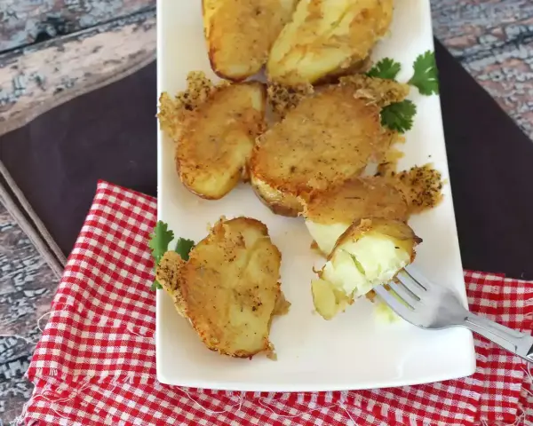 Krispiga, gyllene smashed potatis på en vit tallrik, garnerade med persilja. En gaffel visar en mjuk insida.