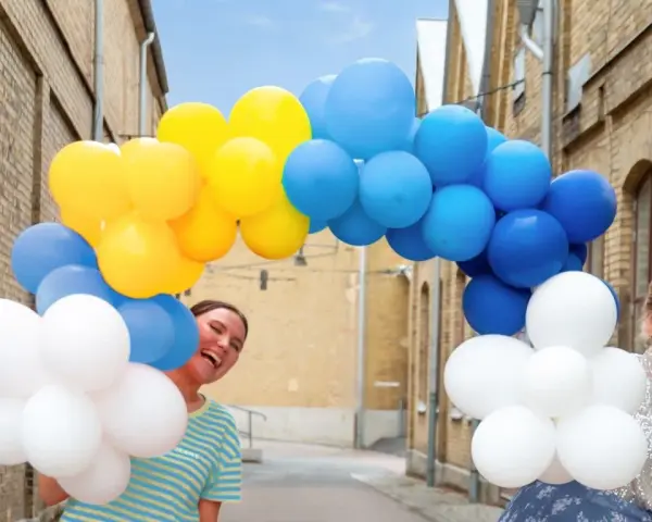 Två glada personer gömmer sig bakom en båge av gula, blå och vita ballonger. Perfekt för studentmottagning lekar!