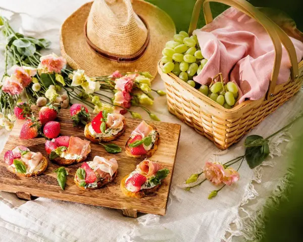 En härlig picknick med bruschetta toppad med jordgubbar och parmaskinka, färska jordgubbar, vindruvor i korg och en stråhatt.