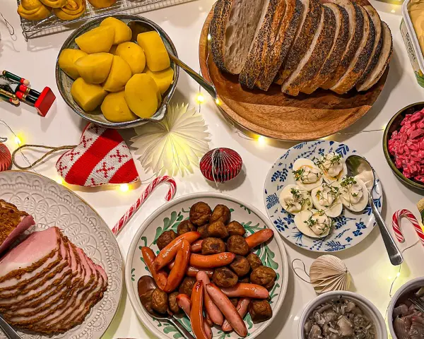 Julbord med jullekar vid bordet: skinka, köttbullar, prinskorv, sill, ägg och bröd. Julpynt och ljusslingor skapar stämning.