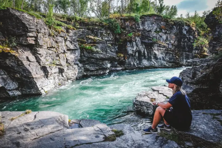 En kvinna sitter vid en forsande turkos flod omgiven av klippor. En plats för unika upplevelser i naturen.