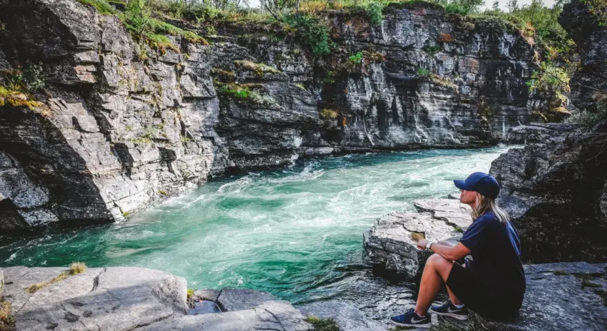 En kvinna sitter vid en forsande turkos flod omgiven av klippor. En plats för unika upplevelser i naturen.