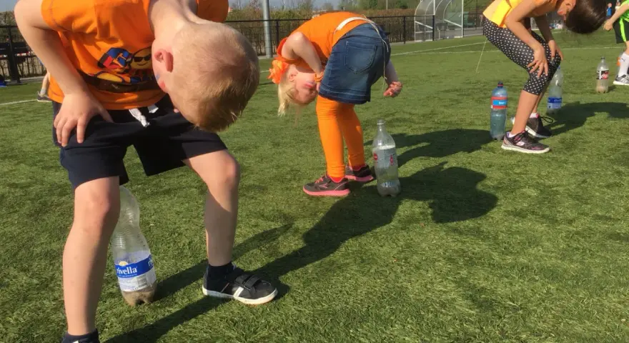 Barn i orange kläder böjer sig framåt för att försöka fånga en flaska med munnen. En del av kalas lekarna.