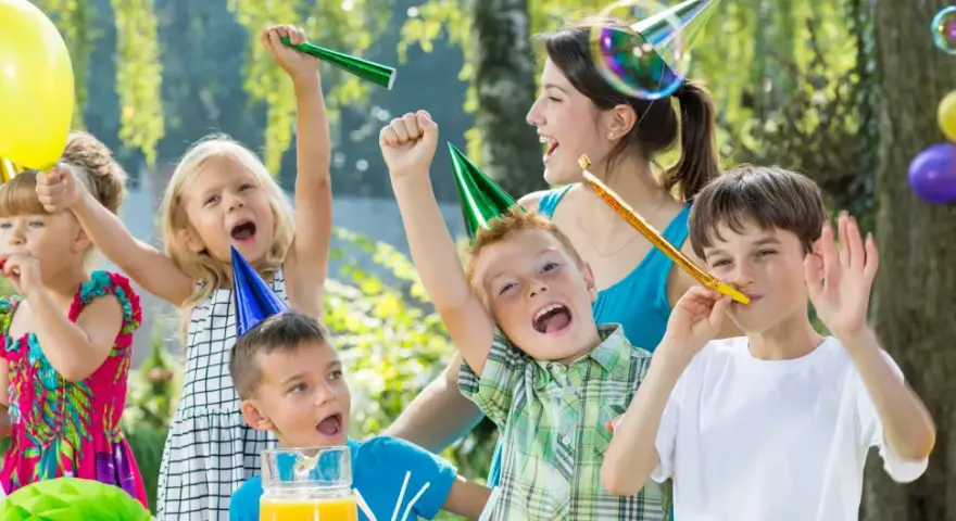 Barn firar ett 7-årskalas utomhus med ballonger, serpentiner och såpbubblor. Glada barn med partyhattar blåser i tutor.