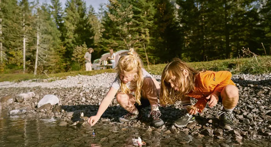 Två barn leker med pappersbåtar vid en sjö, kanske en del av roliga femkampsgrenar. I bakgrunden syns ett tält och träd.