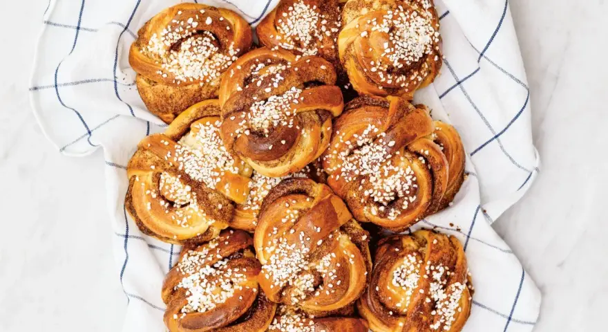 En hög med gyllene kanelbullar, toppade med pärlsocker, ligger på en vit handduk med blå rutor. Camilla Hamid skulle vara stolt över dessa kanelbullar.