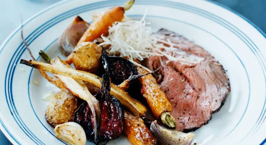 En tallrik med skivad rostbiff, rostade rotfrukter som morötter och palsternacka, samt vitlök. Ett perfekt rostbiff recept för en festmåltid.