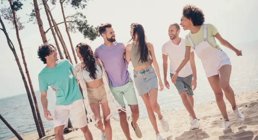 En grupp glada vänner på stranden, en riktig tipspromenad med twist. De skrattar och går längs vattnet bland tallar.