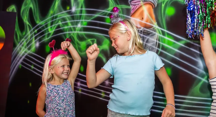 Två glada barn med diadem har roliga lekar inomhus. De dansar framför en neonfärgad bakgrund.