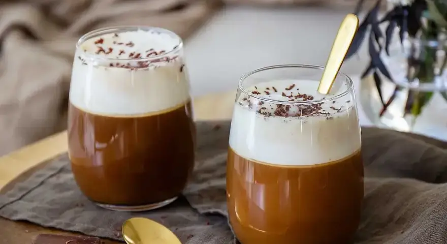 Två glas med krämig kaffe Karlsson, toppad med vispad grädde och chokladströssel. En guldig sked ligger i det ena glaset.