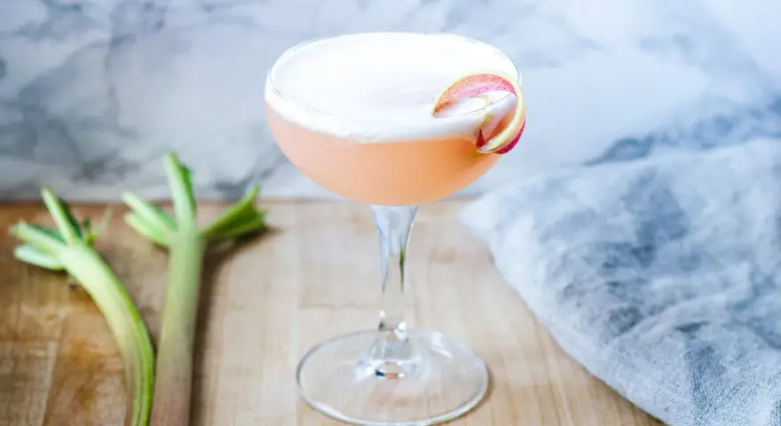En rosa rabarber sour-cocktail med skum och en rabarberdekoration. Två rabarberstjälkar ligger bredvid.