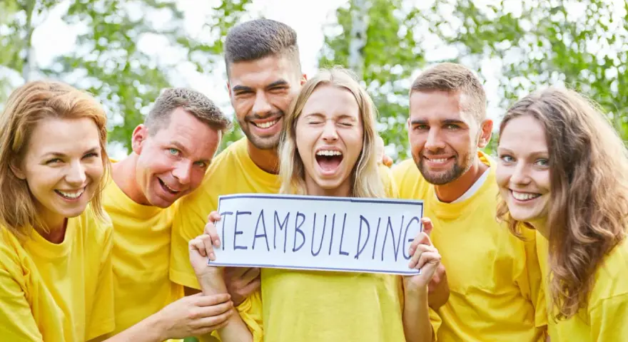 En grupp glada människor i gula tröjor håller upp en skylt med texten "TEAMBUILDING". De ser ut att ha roliga uppdrag i grupp.