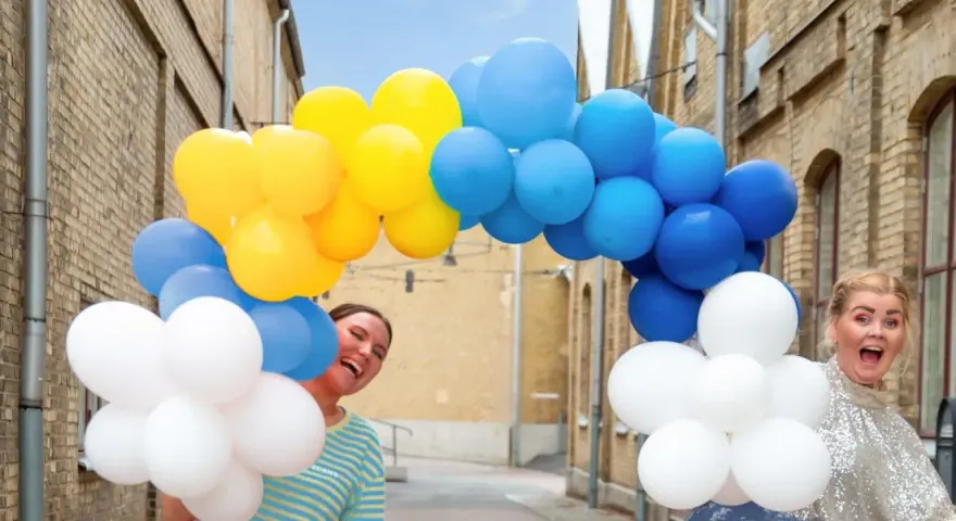 Två glada personer gömmer sig bakom en båge av gula, blå och vita ballonger. Perfekt för studentmottagning lekar!