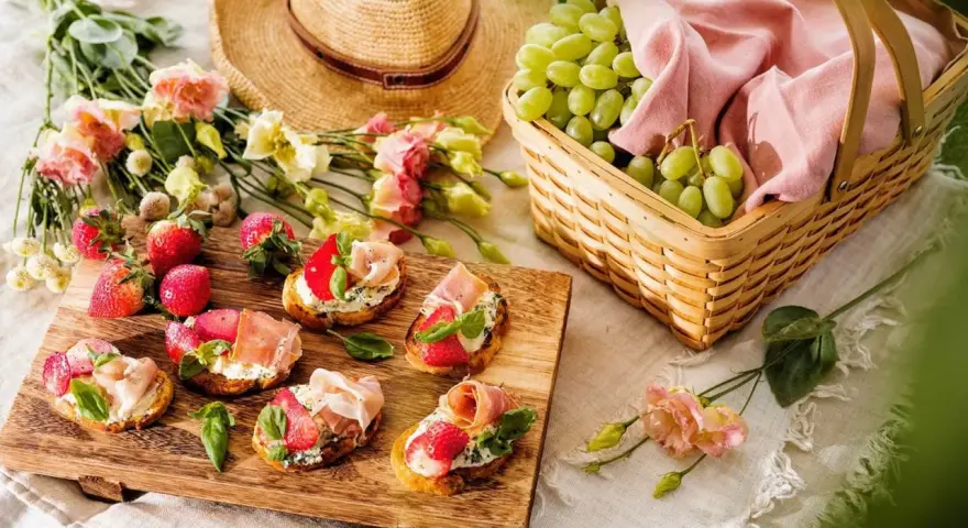 En härlig picknick med bruschetta toppad med jordgubbar och parmaskinka, färska jordgubbar, vindruvor i korg och en stråhatt.
