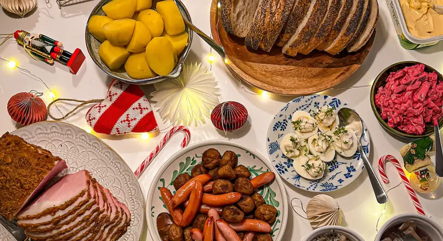Julbord med jullekar vid bordet: skinka, köttbullar, prinskorv, sill, ägg och bröd. Julpynt och ljusslingor skapar stämning.