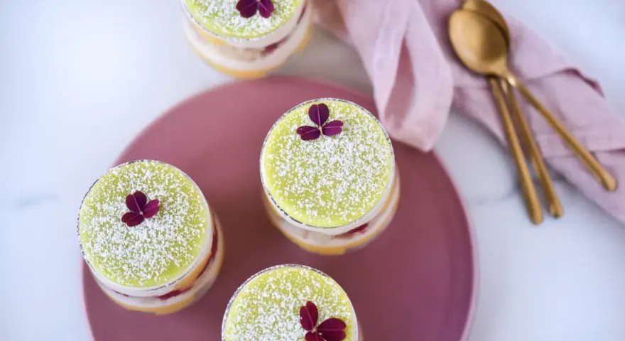 Fyra små prinsesstårtor i glas, dekorerade med florsocker och en lila blomma. En rosa duk och guldfärgade skedar ligger bredvid.