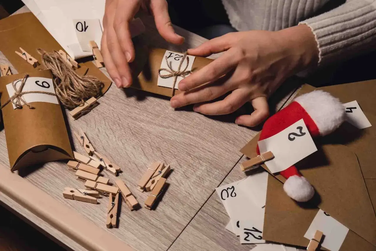 Händer som pysslar med ett julkalender diy. Små bruna paket med siffror och klädnypor ligger utspridda.