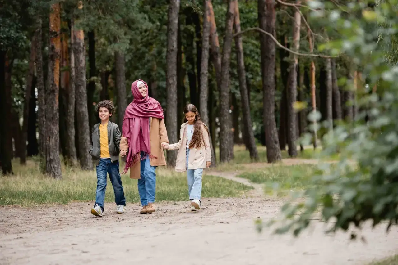 Familj p&aring; en tipspromenad med twist i skogen. En kvinna med hijab och hennes tv&aring; barn g&aring;r hand i hand p&aring; en grusv&auml;g bland h&ouml;ga tr&auml;d.