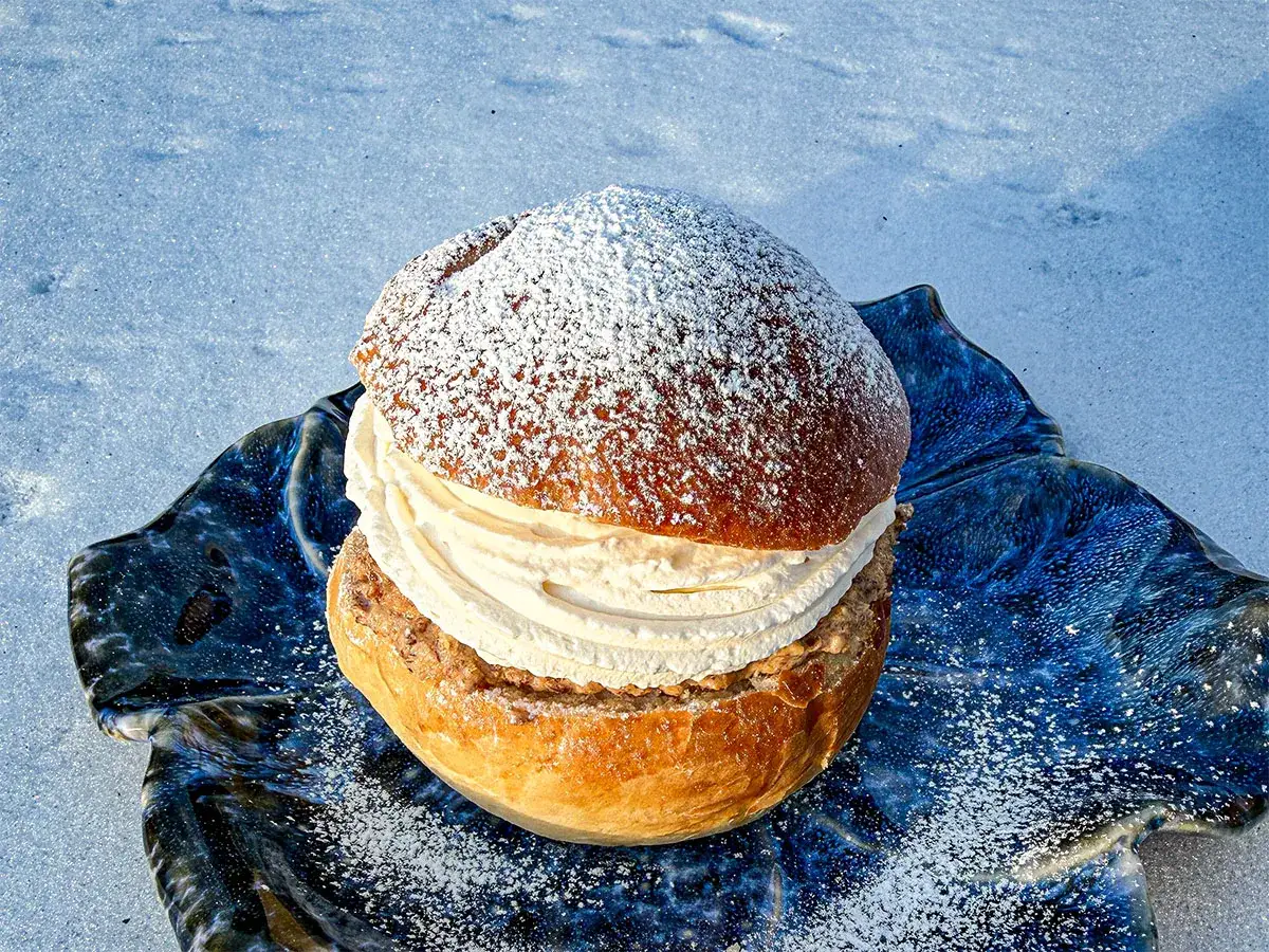 En semla med rikligt med grädde och mandelmassa till semlor, pudrad med florsocker, vilar på en blå, oregelbunden tallrik i snön.