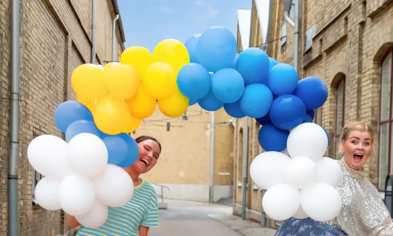 Tv&aring; glada personer g&ouml;mmer sig bakom en b&aring;ge av gula, bl&aring; och vita ballonger. Perfekt f&ouml;r studentmottagning lekar!