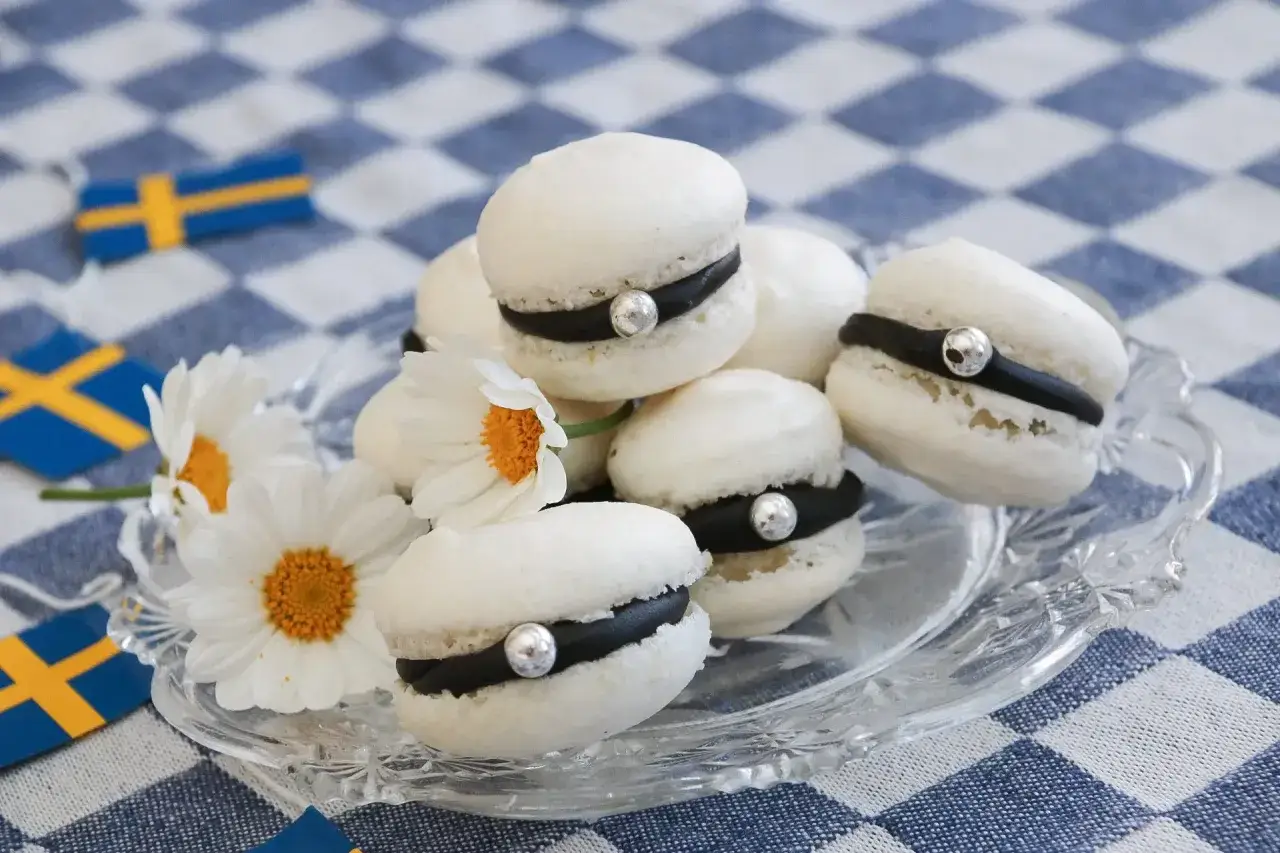 Vita macarons med svart fyllning och silverkulor, dekorerade med pr&auml;stkragar och svenska flaggor. Roliga saker att baka hemma f&ouml;r en festlig stund!