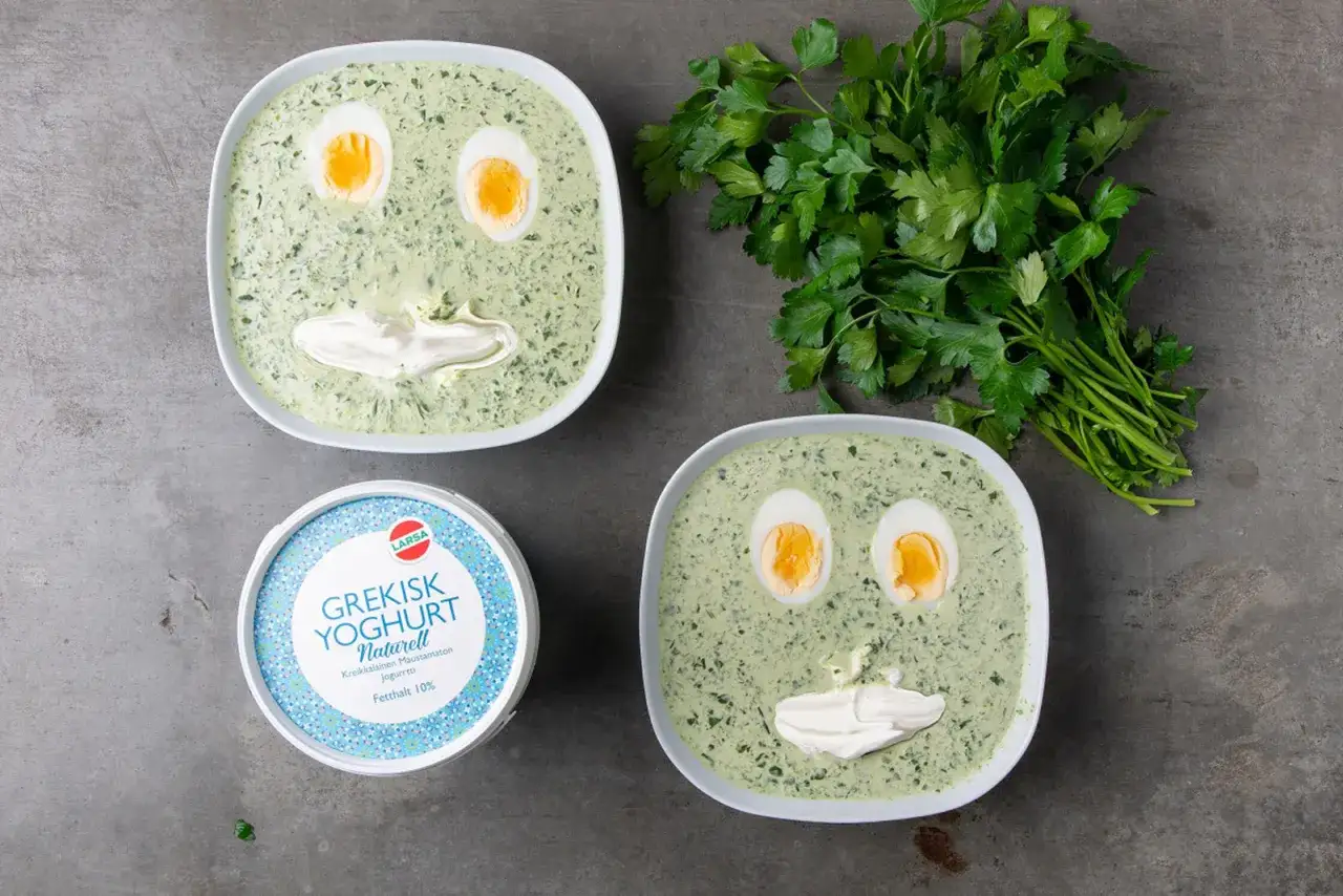 Två skålar med spenatsoppa dekorerade som ansikten, med kokta ägg som ögon och en klick yoghurt som mun. Bredvid ligger en bunt färsk persilja och en burk grekisk yoghurt. Ett roligt spenatsoppa recept!