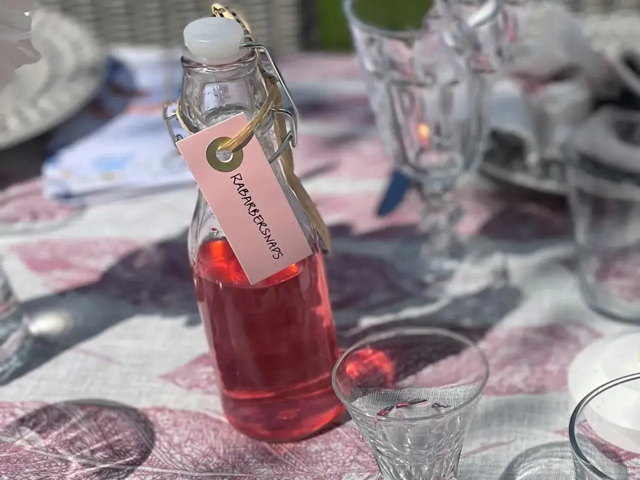 En flaska med rosa rabarbersnaps står på ett dukat bord. Glas och servetter syns i bakgrunden.