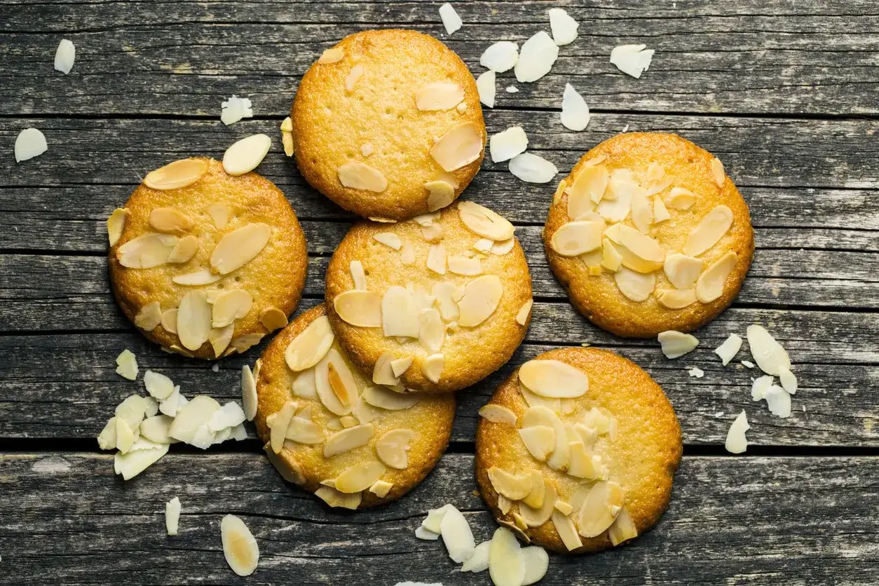 Sju gyllene bondkakor med mandelflagor ligger utspridda på en mörk träyta. Perfekta för ett bondkakor recept.