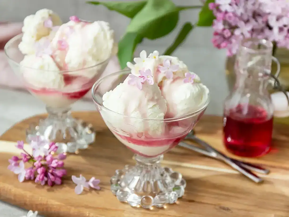 Glassb&auml;gare med vaniljglass, toppad med syrenblommor och r&ouml;d sirap. En flaska med sirap och syrenkvistar i bakgrunden. Ett perfekt syrensirap recept!