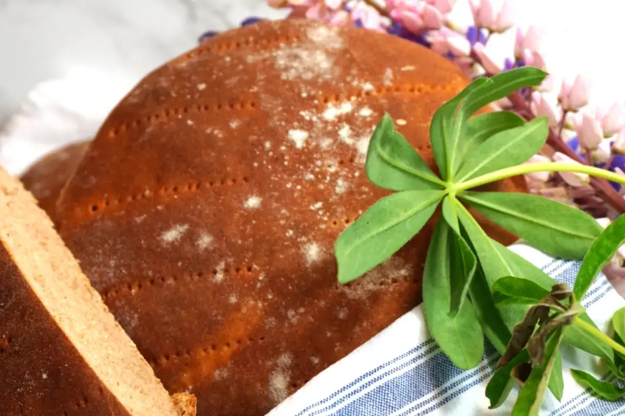 Här är ett härligt rågkakor recept! En rund, mörkbrun rågkaka med prickar och lite mjöl på, bredvid en bit av en annan kaka. Gröna blad och rosa lupiner ger en somrig känsla.