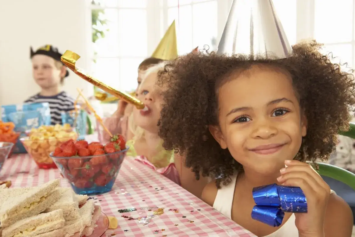 Glada barn med partyhattar och tuta p&aring; ett kalas f&ouml;r en 4-&aring;ring. Jordgubbar, sm&ouml;rg&aring;sar och snacks p&aring; bordet.
