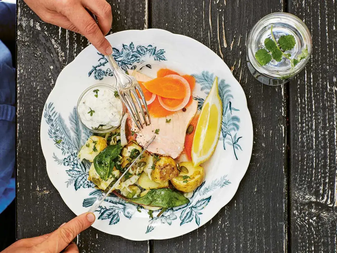 En tallrik med inkokt lax, potatis, grönsaker och en citronskiva. En person skär i laxen med en kniv och gaffel.