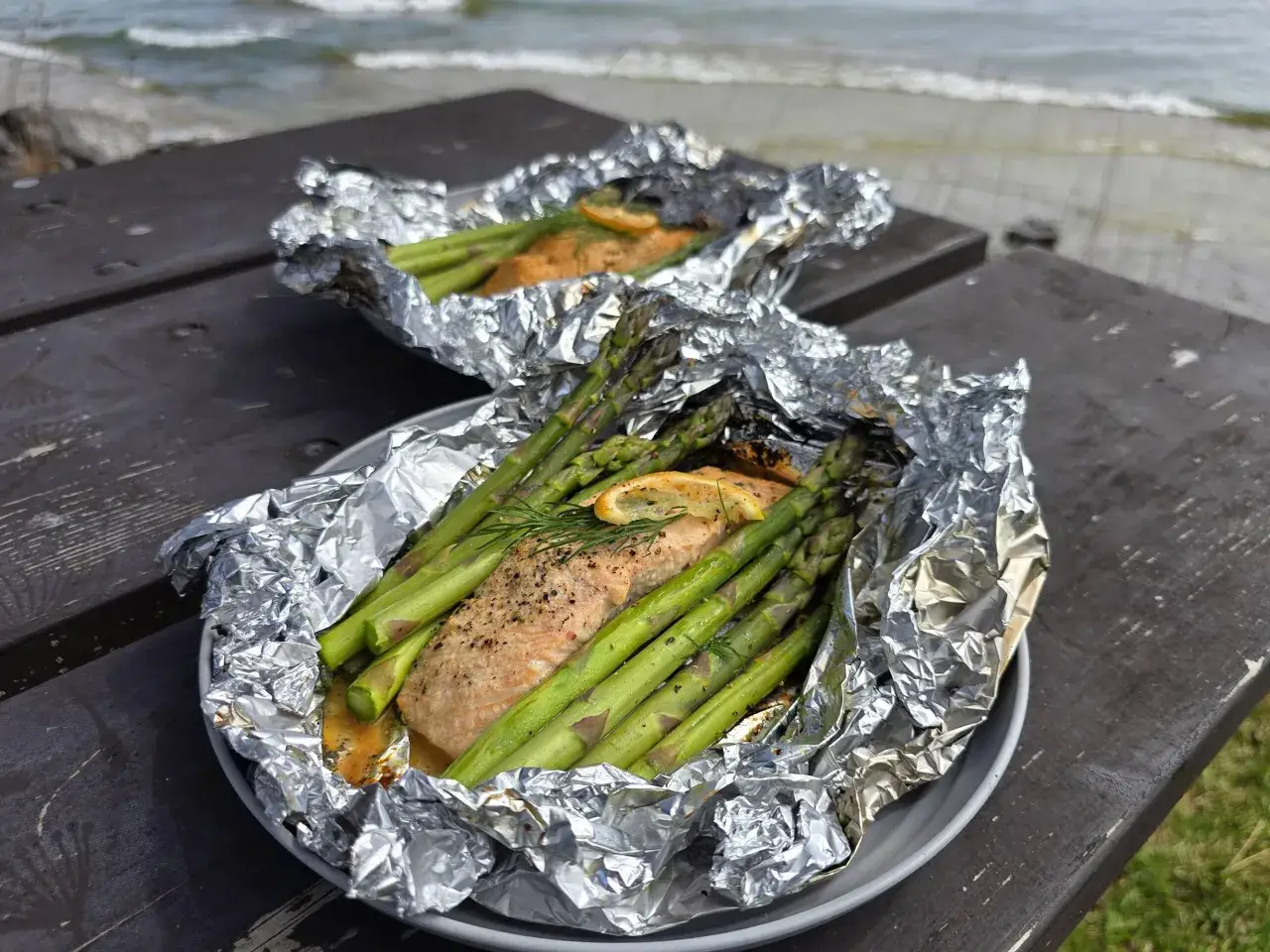 Två fiskpaket med lax och sparris på en grill, redo att avnjutas vid havet.