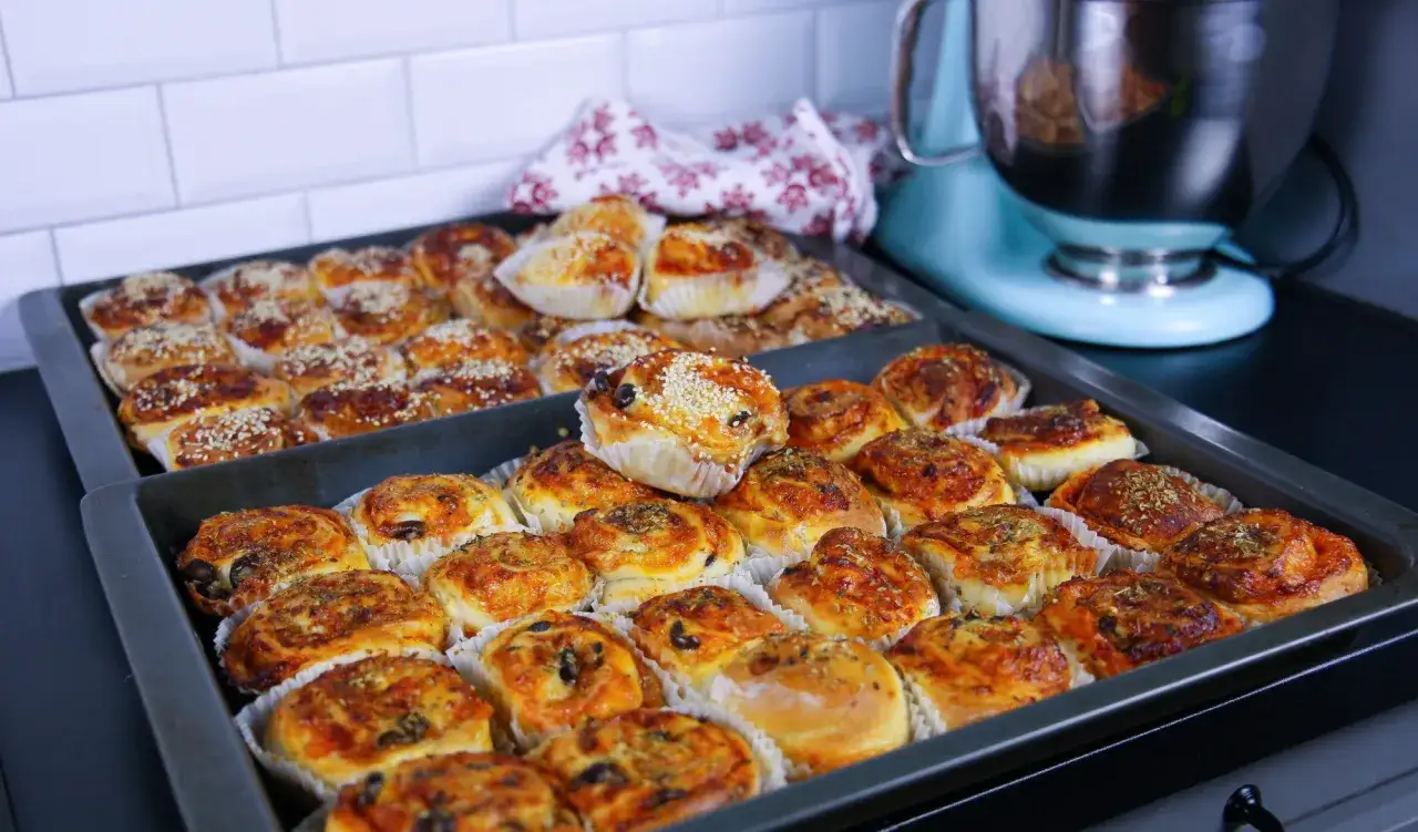 Två plåtar med nybakade pizza bullar, vissa toppade med sesamfrön, andra med örter och ost. En ljusblå köksmaskin syns i bakgrunden.