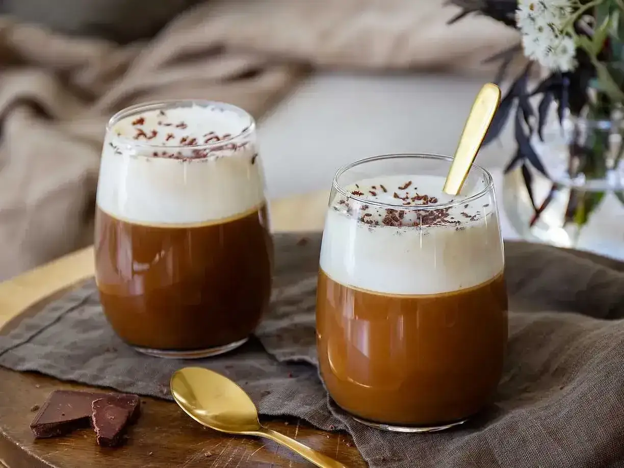 Tv&aring; glas med kr&auml;mig kaffe Karlsson, toppad med vispad gr&auml;dde och chokladstr&ouml;ssel. En gyllene sked ligger i det ena glaset.