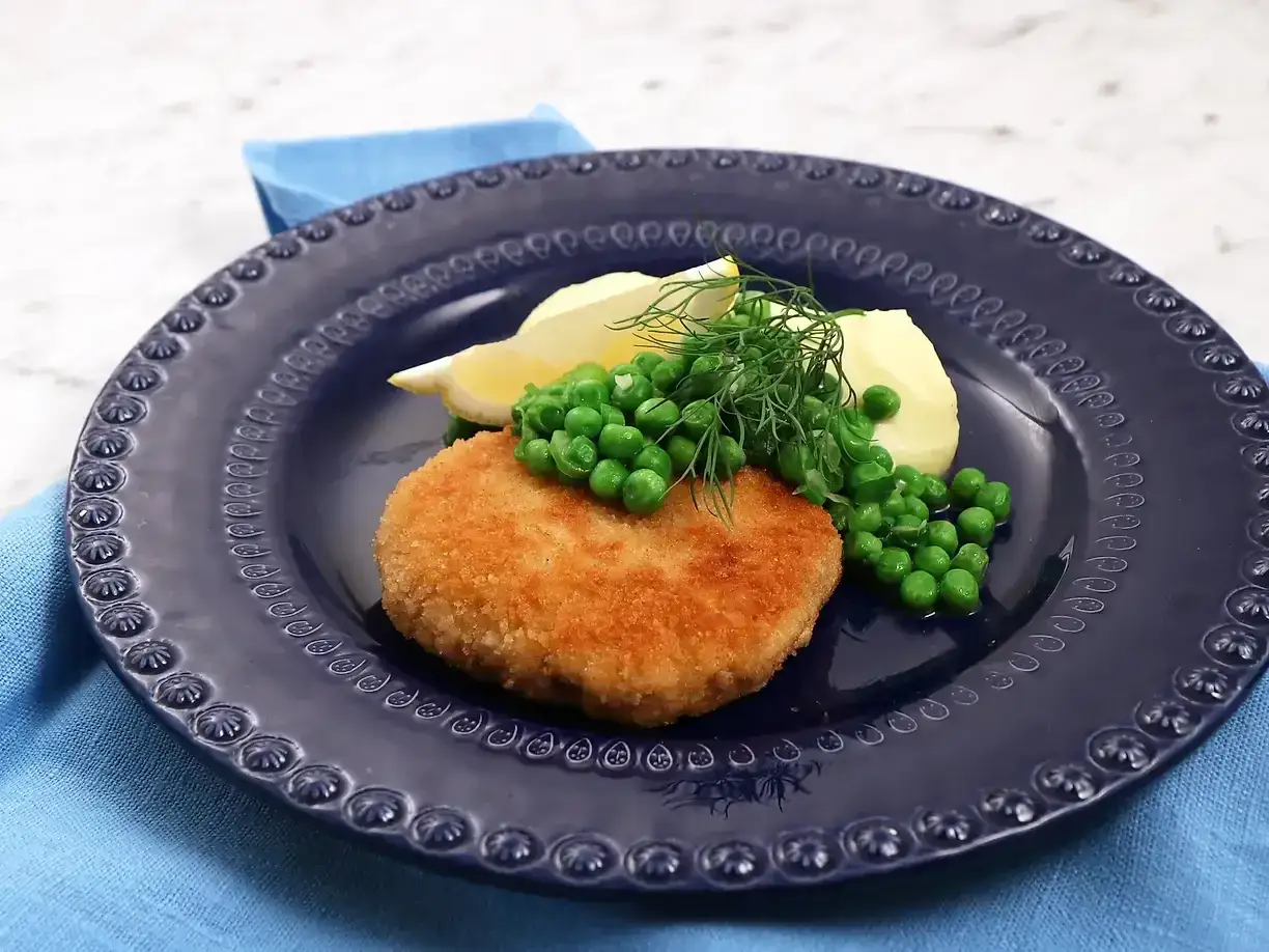 En gyllene havets wallenbergare serveras med potatis, gröna ärtor och citronklyfta på en mörkblå tallrik.